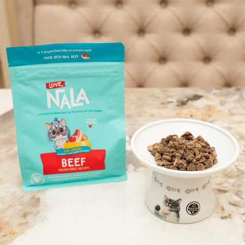 A bag of Love Nala beef grain-free cat food stands next to a white bowl filled with the kibble on a marble countertop, with a tufted leather sofa in the background.