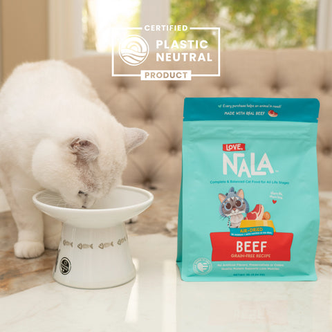 A white cat eating from a white elevated bowl next to a teal and red bag of Nala Love Beef dry cat food, with a 'Certified Plastic Neutral Product' badge overlay at the top.