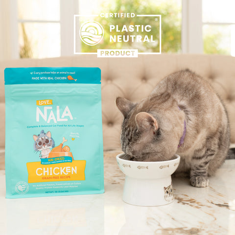 A cat eating from a white elevated bowl next to a bag of Love Nala chicken Organically Raised cat food. A 'Certified Plastic Neutral Product' badge is shown at the top of the image.